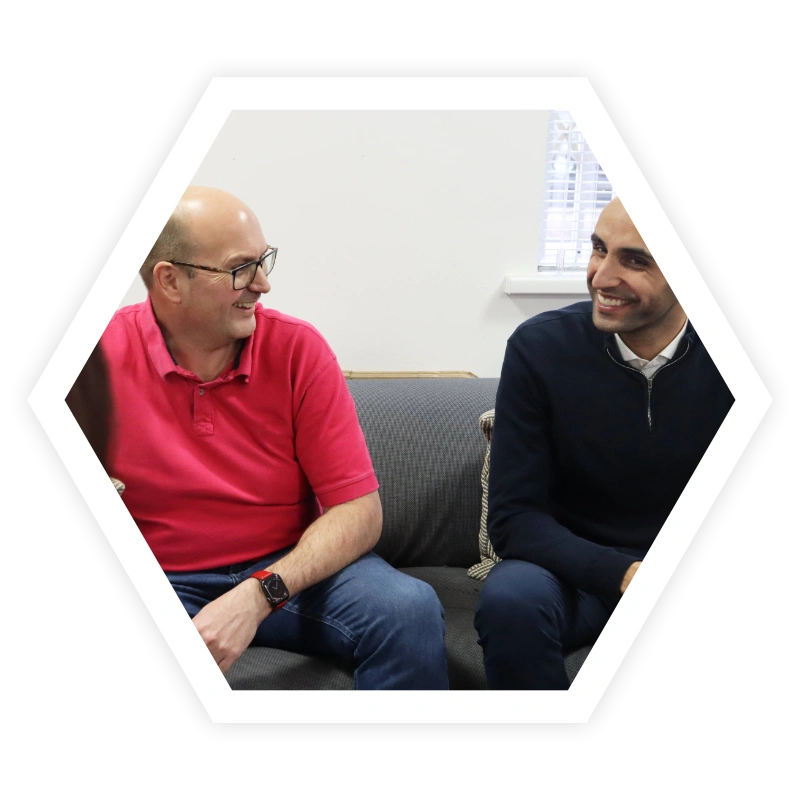 Two men sitting on a couch one wearing red top the other black jumper